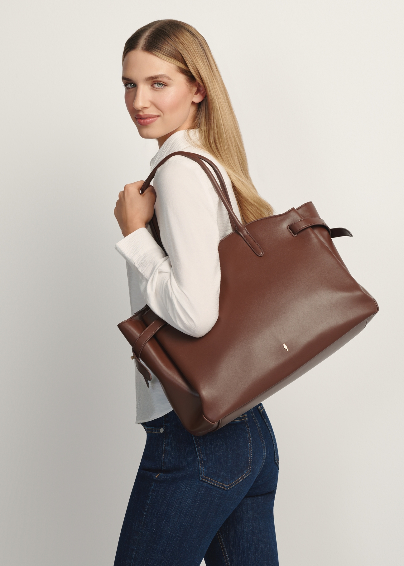 Duża brązowa torebka damska TOREC-1135-1H(W26) - widok produktu z przodu, na modelce stojącej bokiem, na jasnym tle. Widoczna połowa sylwetki. 