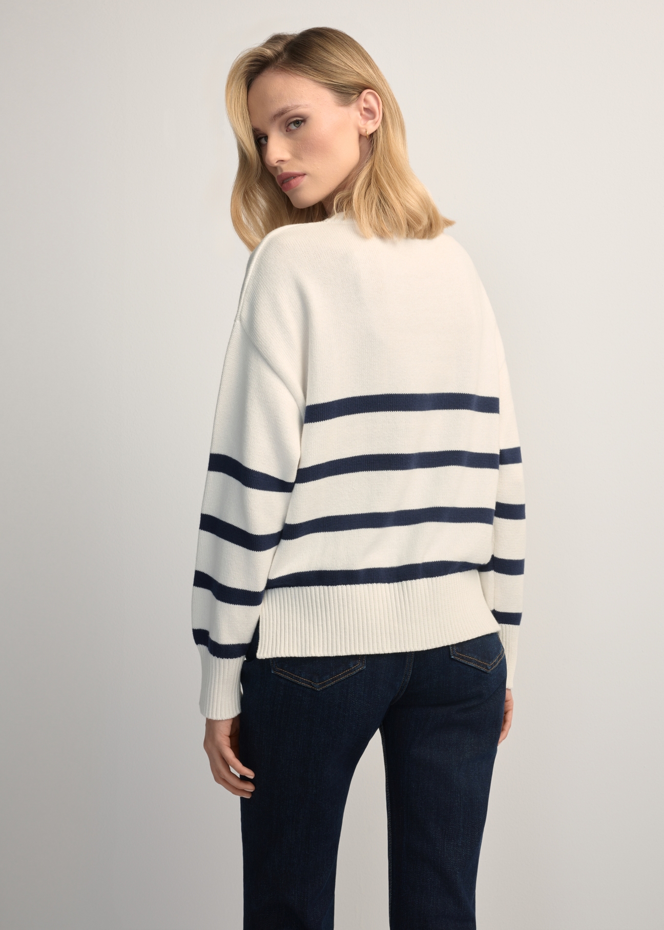 Kremowy sweter damski w paski SWEDT-0258-0B(W26) - widok produktu z tyłu, na modelce, na jasnym tle. Widoczna sylwetka do kolan. 